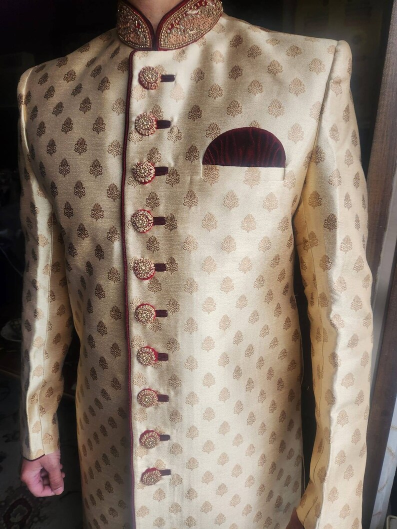 May include: A gold and maroon men's formal jacket with a button-up closure and a small pocket on the left side. The jacket has a floral pattern and is decorated with maroon buttons.