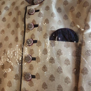 May include: A beige men's garment with a maroon collar and intricate gold embroidery. The garment has a row of decorative buttons with rhinestones and a maroon pocket on the left side.