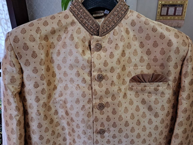 May include: A beige men's jacket with a brown patterned design and a brown velvet pocket. The jacket has a high collar with a brown beaded trim.