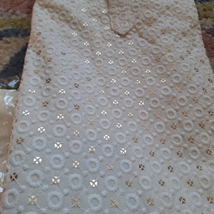 Puede incluir: Tela blanca con un delicado patr&oacute;n de peque&ntilde;os c&iacute;rculos y cruces, bordada con hilo dorado.