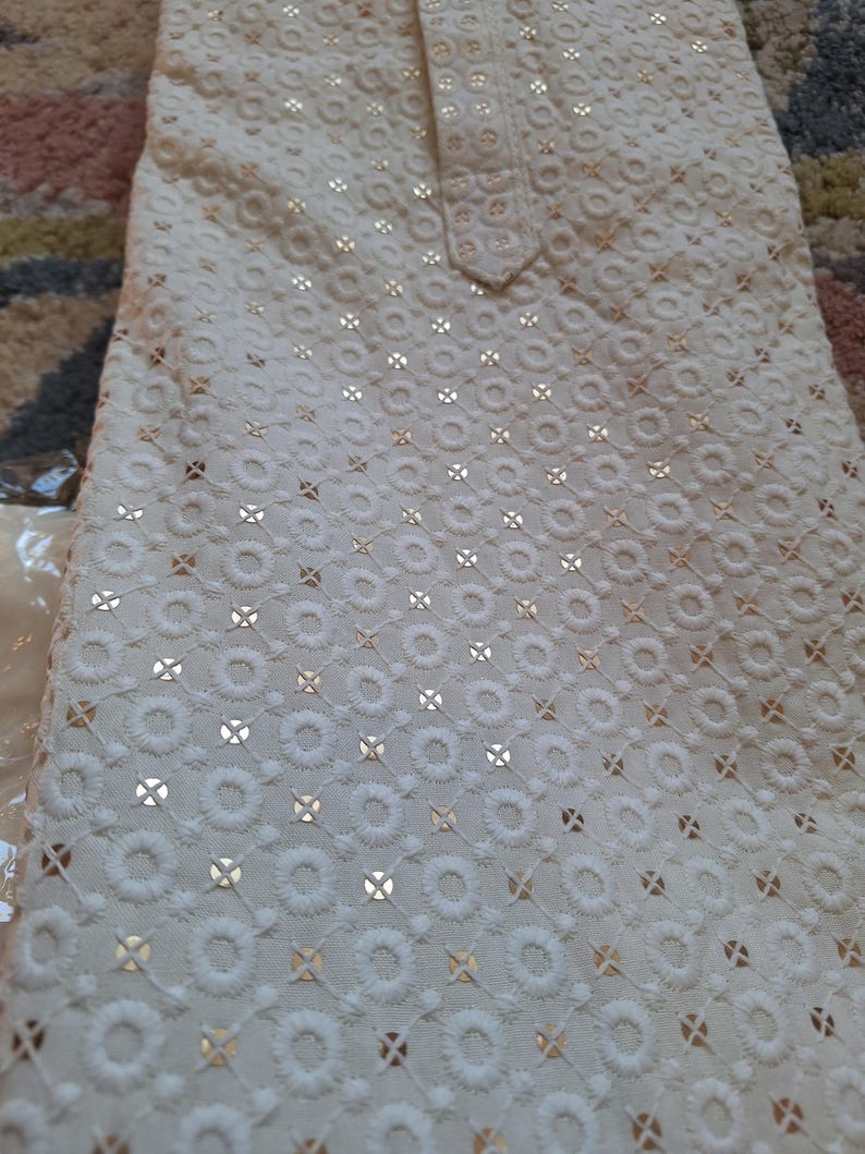 Puede incluir: Tela blanca con un delicado patr&oacute;n de peque&ntilde;os c&iacute;rculos y cruces, bordada con hilo dorado.