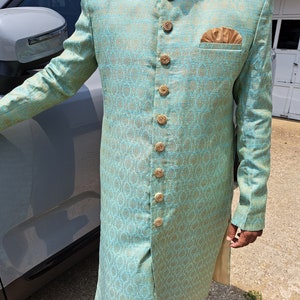 Puede incluir: Un sherwani de brocado azul claro y dorado con botones dorados ornamentados y un pa&ntilde;uelo de bolsillo a juego. La prenda tradicional india presenta un cuello mandar&iacute;n y patrones tejidos intrincados, adecuada para ocasiones formales.