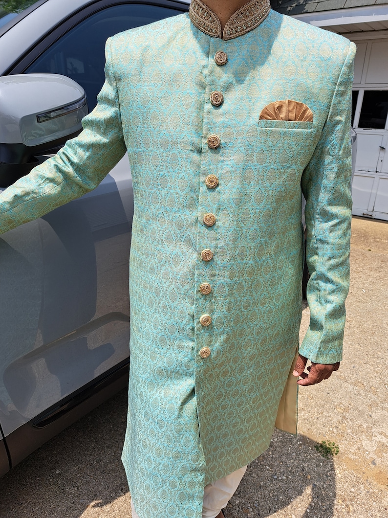 Puede incluir: Un sherwani de brocado azul claro y dorado con botones dorados ornamentados y un pa&ntilde;uelo de bolsillo a juego. La prenda tradicional india presenta un cuello mandar&iacute;n y patrones tejidos intrincados, adecuada para ocasiones formales.