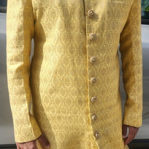 K&ouml;nnte beinhalten: Eine goldgelbe Jacke mit langen &Auml;rmeln, aufwendigen, sich wiederholenden Mustern und dekorativen Kn&ouml;pfen. Die Jacke hat einen Stehkragen mit goldener Stickerei und ein Einstecktuch. Das Outfit wird mit einer wei&szlig;en Hose kombiniert. Festliche Kleidung.