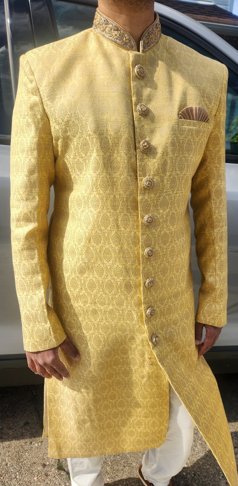 K&ouml;nnte beinhalten: Eine goldgelbe Jacke mit langen &Auml;rmeln, aufwendigen, sich wiederholenden Mustern und dekorativen Kn&ouml;pfen. Die Jacke hat einen Stehkragen mit goldener Stickerei und ein Einstecktuch. Das Outfit wird mit einer wei&szlig;en Hose kombiniert. Festliche Kleidung.