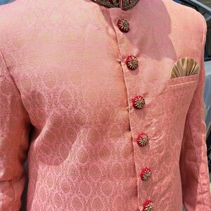 Puede incluir: Chaqueta de brocado rosa con cuello mandar&iacute;n y detalles dorados y rojos ornamentados. La chaqueta presenta botones decorativos y un pa&ntilde;uelo de bolsillo. La tela tiene un sutil estampado floral.