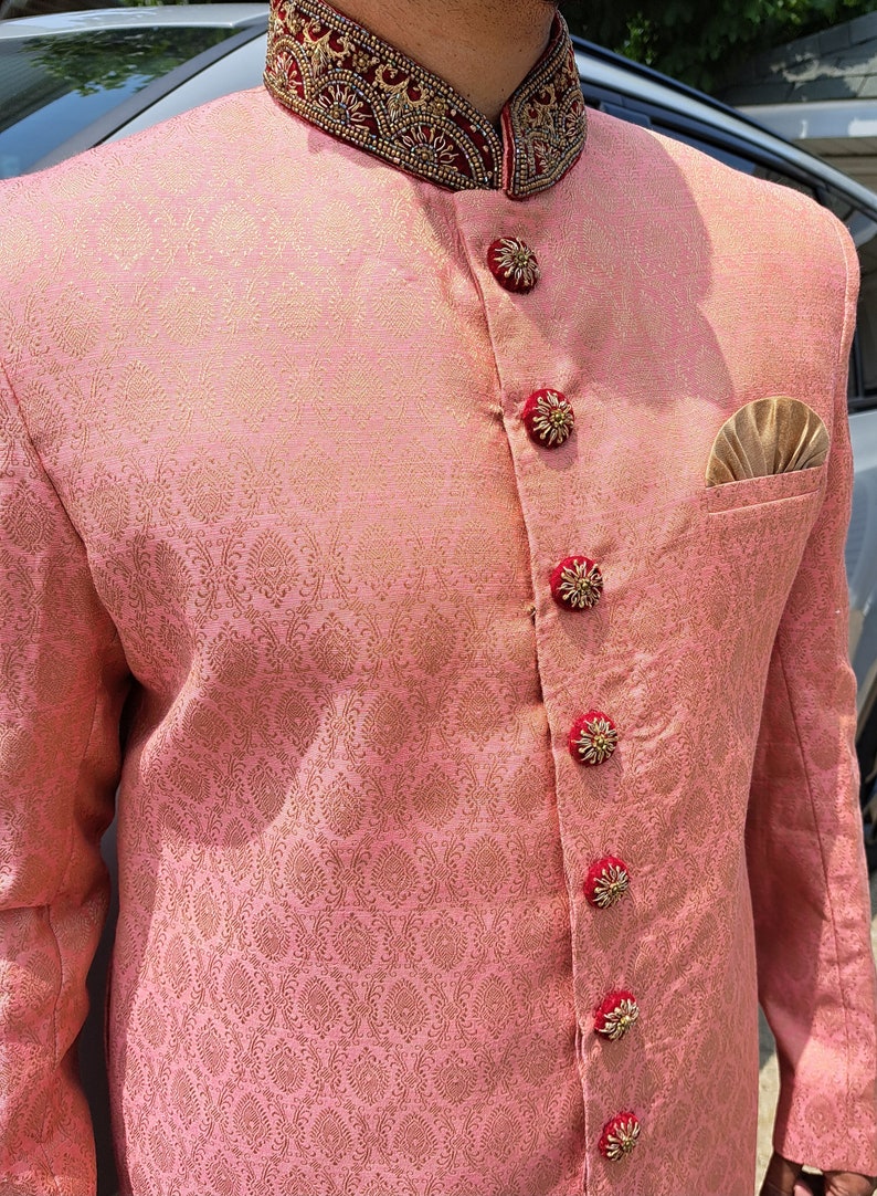 Puede incluir: Chaqueta de brocado rosa con cuello mandar&iacute;n y detalles dorados y rojos ornamentados. La chaqueta presenta botones decorativos y un pa&ntilde;uelo de bolsillo. La tela tiene un sutil estampado floral.