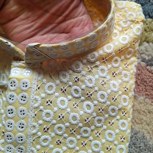 May include: A yellow fabric with white embroidered circles and a criss-cross pattern. The fabric has a gold trim around the edge.