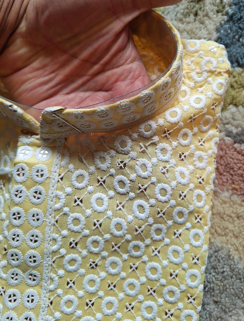 May include: A yellow fabric with white embroidered circles and a criss-cross pattern. The fabric has a gold trim around the edge.