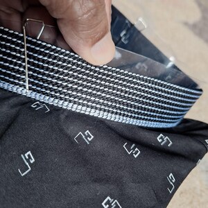 May include: Close-up of a black fabric with a white geometric pattern and a contrasting white and black striped band. The fabric has a repeating white geometric design. A metal clip is attached to the fabric.