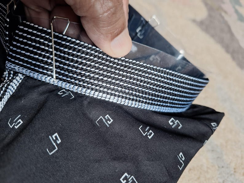 May include: Close-up of a black fabric with a white geometric pattern and a contrasting white and black striped band. The fabric has a repeating white geometric design. A metal clip is attached to the fabric.