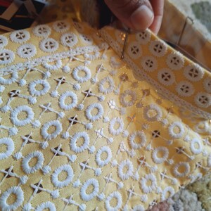 May include: A yellow fabric with white embroidered circles and a white lace trim. The fabric is being held by a hand.