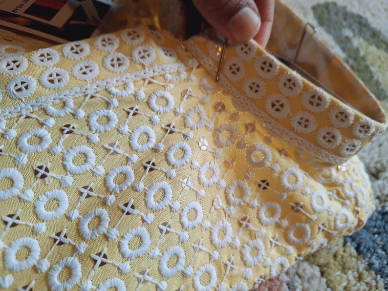 May include: A yellow fabric with white embroidered circles and a white lace trim. The fabric is being held by a hand.