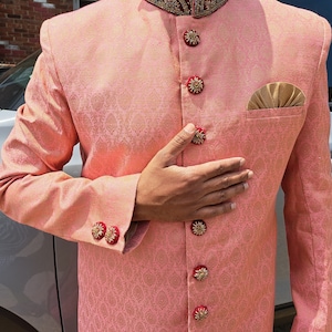Puede incluir: Chaqueta melocot&oacute;n estampada con botones y detalles de pu&ntilde;os dorados y rojos ornamentados. La chaqueta tiene un bolsillo con un pa&ntilde;uelo dorado. El cuello tiene un dise&ntilde;o bordado en rojo oscuro y dorado. Ropa formal.