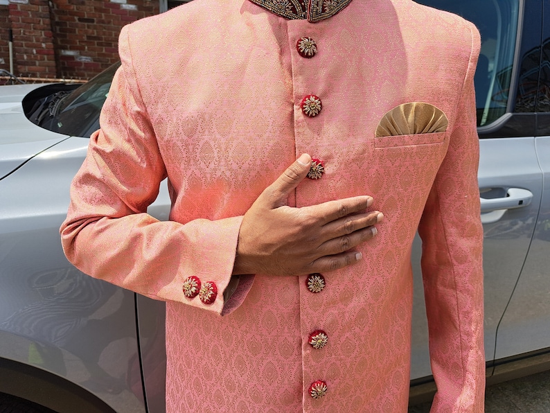 Puede incluir: Chaqueta melocot&oacute;n estampada con botones y detalles de pu&ntilde;os dorados y rojos ornamentados. La chaqueta tiene un bolsillo con un pa&ntilde;uelo dorado. El cuello tiene un dise&ntilde;o bordado en rojo oscuro y dorado. Ropa formal.
