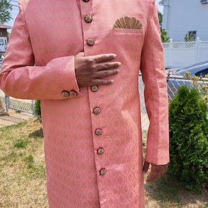 Puede incluir: Un sherwani rosa estampado con detalles dorados y rojos ornamentados. El abrigo largo presenta botones decorativos y un pa&ntilde;uelo de bolsillo. El cuello y los pu&ntilde;os est&aacute;n adornados con intrincados bordados. Es una prenda formal tradicional del sur de Asia.