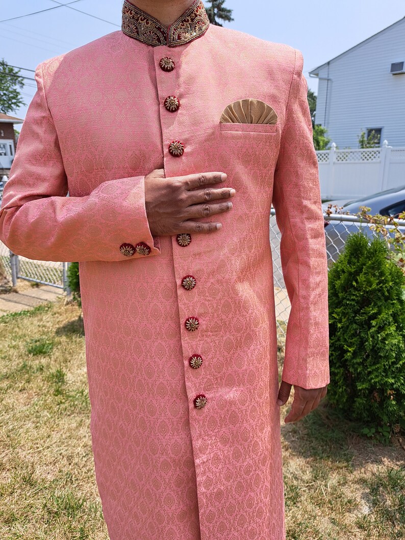 Puede incluir: Un sherwani rosa estampado con detalles dorados y rojos ornamentados. El abrigo largo presenta botones decorativos y un pa&ntilde;uelo de bolsillo. El cuello y los pu&ntilde;os est&aacute;n adornados con intrincados bordados. Es una prenda formal tradicional del sur de Asia.