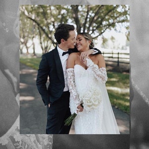 May include: A bride and groom are smiling at each other. The bride is wearing a white lace wedding dress with off-the-shoulder sleeves and a bouquet of white roses. The groom is wearing a black tuxedo with a black bow tie.