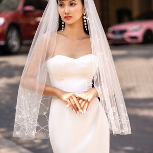 Può includere: Una donna che indossa un abito da sposa bianco senza spalline con un lungo velo decorato con piccoli cristalli. Il velo è trasparente e presenta un delicato motivo floreale. La donna è in piedi davanti a un edificio con un'auto rosa sullo sfondo.