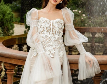 May include: A white bridal gown with a lace bodice and tulle skirt. The gown has long, puffed sleeves with lace detailing. The model is standing in front of a fountain with a stone railing.