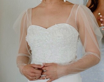 May include: A white wedding dress with a sweetheart neckline and long sleeves. The sleeves are made of sheer tulle and have a puffed detail at the shoulder. The bodice is covered in delicate beading.