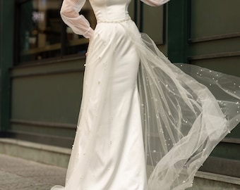 Puede incluir: Un vestido de novia blanco con una superposición de encaje transparente y una cola de tul con cuentas. El vestido tiene un escote de corazón y un corpiño ajustado. La modelo lleva el vestido con un bolero de encaje blanco.
