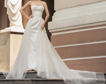 Puede incluir: Una mujer con un vestido de novia blanco sin tirantes y una larga cola se encuentra de pie en una escalera frente a un edificio. El vestido tiene un corpiño ajustado y una falda fluida. La mujer sonríe y mira a la cámara.