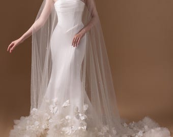 Puede incluir: Un vestido de novia blanco sin tirantes con silueta de sirena. El vestido presenta una falda en capas con aplicaciones florales y una capa transparente con cuentas. La modelo lleva pendientes de perlas.