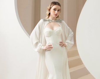 May include: A white bridal gown with a fitted bodice and a flowing skirt. The gown features a sheer cape with a beaded neckline. The model is standing in a white room with a circular archway.