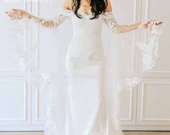 May include: A white off-the-shoulder bridal gown with a fitted bodice and a flowing skirt. The gown features delicate lace detailing on the sleeves and hem. The gown is paired with a sheer lace overlay that extends from the shoulders to the floor.