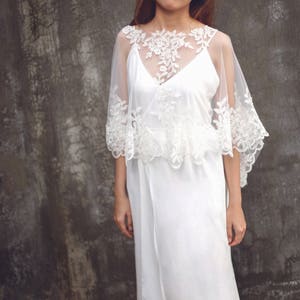 May include: A white lace bridal cape with a delicate floral pattern. The cape is sheer and has a scalloped edge. It is worn over a white dress.