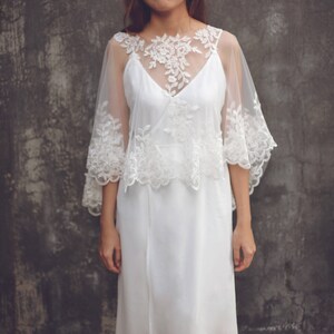 May include: A white lace overlay with a delicate floral pattern. The overlay is sheer and has a scalloped edge. It is worn over a white dress.