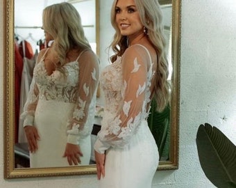 May include: A white wedding dress with a fitted bodice and a flowing skirt. The dress features a sheer lace overlay with delicate floral embroidery and long sleeves. The bride is standing in front of a mirror, looking at her reflection.