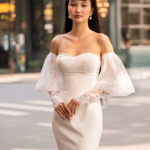 May include: A white off-the-shoulder bridal gown with long, puffy sleeves and a fitted bodice. The gown has a mermaid silhouette and a lace trim on the sleeves.