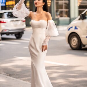 May include: A white off-the-shoulder bridal gown with a fitted bodice and a flowing skirt. The gown features delicate lace sleeves with puffed shoulders. The model is standing on a sidewalk in front of a building.