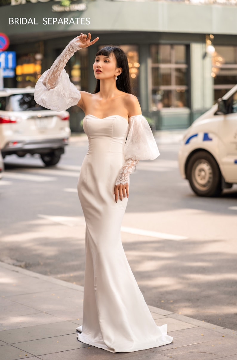 May include: A white off-the-shoulder bridal gown with a fitted bodice and a flowing skirt. The gown features delicate lace sleeves with puffed shoulders. The model is standing on a sidewalk in front of a building.