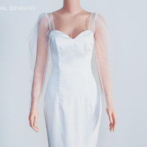 May include: A white bridal dress with a sweetheart neckline and long sheer sleeves. The dress is fitted and has a simple design.