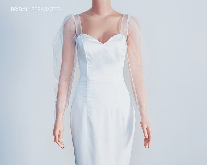 May include: A white bridal dress with a sweetheart neckline and long sheer sleeves. The dress is fitted and has a simple design.
