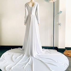 May include: A white long-sleeved gown with a lace-up back and beaded detail at the neckline. The gown has a flowing skirt and a train.