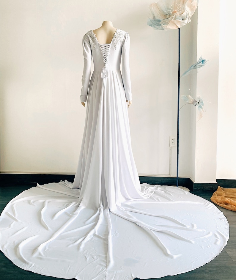 May include: A white long-sleeved gown with a lace-up back and beaded detail at the neckline. The gown has a flowing skirt and a train.