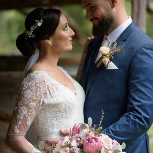 Pu&ograve; includere: Una sposa e uno sposo stanno in piedi vicini, guardandosi negli occhi. La sposa indossa un abito bianco di pizzo e un accessorio per capelli floreale. Lo sposo indossa un abito blu scuro e una camicia bianca. Tengono un bouquet di fiori rosa e bianchi.
