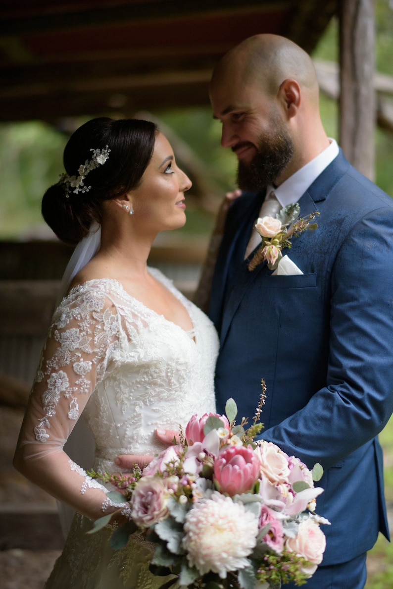 Pu&ograve; includere: Una sposa e uno sposo stanno in piedi vicini, guardandosi negli occhi. La sposa indossa un abito bianco di pizzo e un accessorio per capelli floreale. Lo sposo indossa un abito blu scuro e una camicia bianca. Tengono un bouquet di fiori rosa e bianchi.