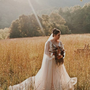 May include: A bride wearing a white wedding dress with a long veil stands in a field of tall grass. She is holding a bouquet of dried flowers in shades of brown, orange, and white. The sun is shining brightly behind her, creating a warm glow.