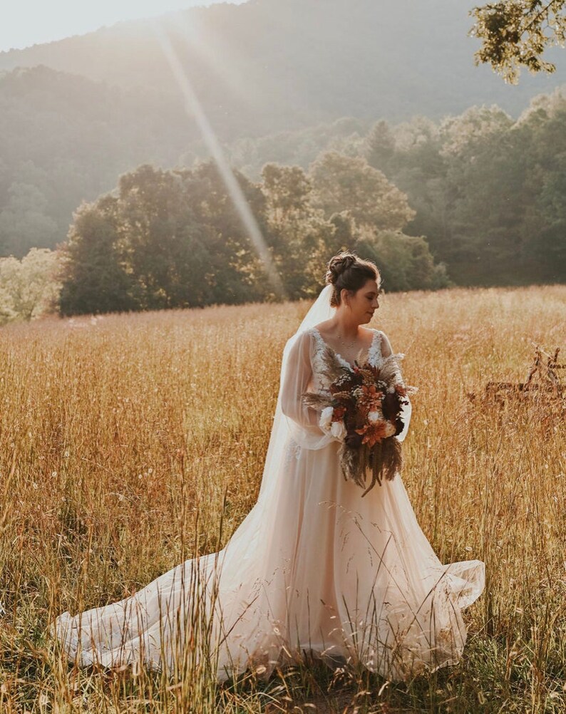 May include: A bride wearing a white wedding dress with a long veil stands in a field of tall grass. She is holding a bouquet of dried flowers in shades of brown, orange, and white. The sun is shining brightly behind her, creating a warm glow.