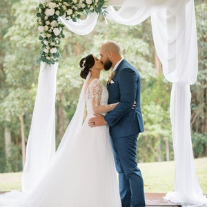 Pu&ograve; includere: Una sposa e uno sposo si baciano sotto un arco di matrimonio bianco decorato con fiori bianchi e verde. L'arco &egrave; drappeggiato con tessuto bianco. La sposa indossa un abito da sposa bianco e lo sposo indossa un abito blu.