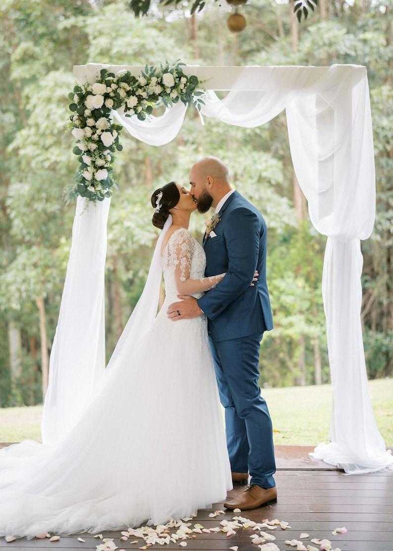 Pu&ograve; includere: Una sposa e uno sposo si baciano sotto un arco di matrimonio bianco decorato con fiori bianchi e verde. L'arco &egrave; drappeggiato con tessuto bianco. La sposa indossa un abito da sposa bianco e lo sposo indossa un abito blu.