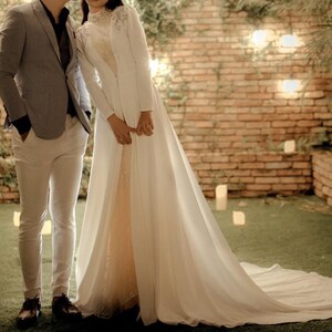 May include: A couple in a wedding setting. The bride is wearing a white gown with a long train and a white jacket. The groom is wearing a gray suit and white pants. They are standing in front of a brick wall with greenery and lights.