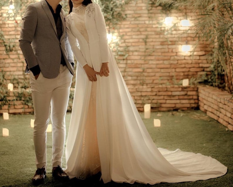 May include: A couple in a wedding setting. The bride is wearing a white gown with a long train and a white jacket. The groom is wearing a gray suit and white pants. They are standing in front of a brick wall with greenery and lights.
