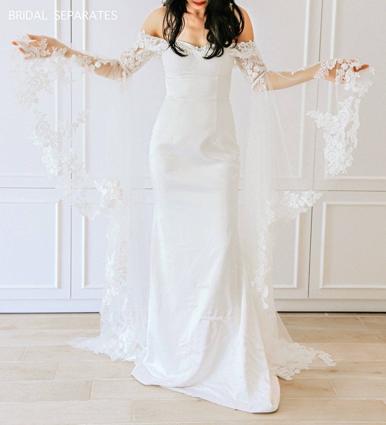 May include: A white off-the-shoulder bridal gown with a fitted bodice and a flowing skirt. The gown features delicate lace detailing on the sleeves and hem. The gown is paired with a sheer lace overlay that extends from the shoulders to the floor.