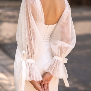 May include: A white bridal gown with a fitted bodice and a flowing skirt. The gown features long, sheer sleeves with delicate lace detailing and a bow at the wrist.